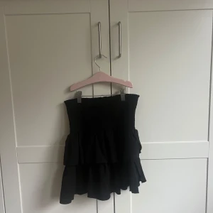 Svart volangkjol - Snygg svart kortkjol med volanger från Neo Noir. Perfekt för en trendig look. Kjolen har en tight passform i midjan och faller fint med sina lager av volanger. En liten rem i kjolen som är lite sönder men annars som ny. Köptes för 450kr och säljer för 100kr.