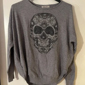 Tunnstickad tröja med nitar onesize - Snygg grå tröja med ett stort, detaljerat dödskallemotiv i glittrande stenar på framsidan. Tröjan har en avslappnad passform och långa ärmar, perfekt för en cool och avslappnad stil. Helt ny. Storlek onesize.