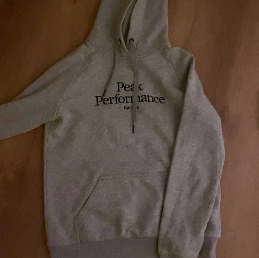 Säljer en stilren grå hoodie från Peak Performance med tryck på bröstet. Den har en klassisk känguruficka och justerbar huva med dragsko. Perfekt för en avslappnad stil.. Neuletakit & Villapaidat.