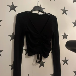 Svart ribbad tröja från Missguided - Snygg svart ribbad tröja från Missguided med v-ringning och knytning framtill. Tröjan har långa ärmar och en croppad passform, perfekt för en trendig look. Passar bra till både jeans och kjol!