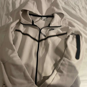 Vit huvtröja från Nike - Säljer en vit huvtröja från Nike med svarta detaljer och dragkedja. Tröjan har en praktisk ficka på ärmen och en justerbar huva. Perfekt för en sportig look eller avslappnade dagar. 