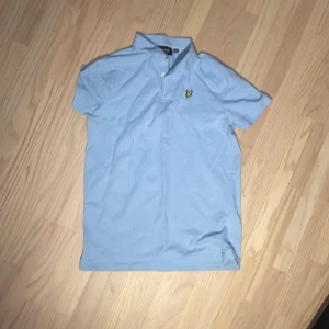 Ljusblå pikétröja från Lyle & Scott - Säljer en ljusblå pikétröja från Lyle & Scott med klassisk krage och knappslå. Tröjan har en broderad logotyp på bröstet i gult och svart. Perfekt för en stilren och avslappnad look.