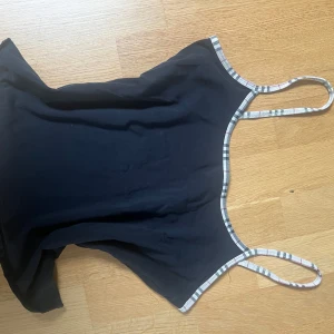 Svart linnetopp från Burberry - Snygg svart linnetopp från Burberry med smala axelband och klassiskt rutigt kantband. Perfekt för en stilren look. Nästan 100% bomull och liten i storleken så passar S