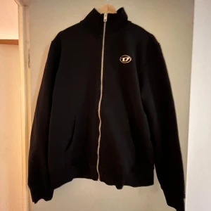 Svart hoodie från Diesel - Säljer en svart hoodie från Diesel med dragkedja och en liten logga på bröstet. Tröjan har långa ärmar och en hög krage. Perfekt för en stilren och bekväm look.