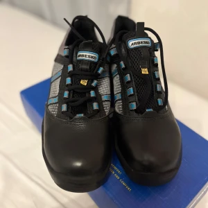 Svarta arbetsskor från Arbesko - Säljer ett par svarta arbetsskor från Arbesko med grå och blå detaljer. Skorna har snörning och är designade för komfort och säkerhet. Perfekta för arbetsmiljöer där extra skydd behövs.