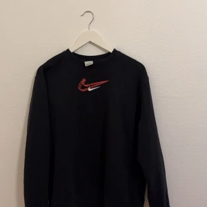 Svart sweatshirt från Nike - Säljer en svart sweatshirt från Nike med det klassiska logotypen i rött och vitt på bröstet. Tröjan har långa ärmar och en rund halsringning. Perfekt för en avslappnad stil.