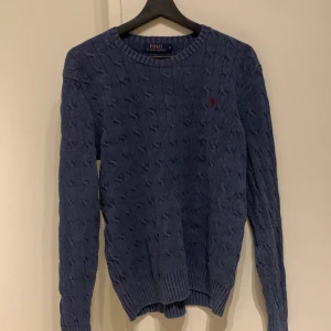 Blå stickad tröja från Polo Ralph Lauren - Säljer en snygg blå stickad tröja från Polo Ralph Lauren med kabelstickat mönster. Tröjan har en liten röd broderad logga på bröstet och långa ärmar. Perfekt för kyligare dagar! Tröjan kan vara lite urtvättad.