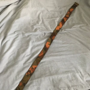 Kamouflagefärgat bälte från J. Lindeberg - Säljer ett coolt kamouflagefärgat bälte från J. Lindeberg, tillverkat i äkta läder. Bältet har en mix av gröna och orange nyanser och är gjort i Italien. Perfekt för att ge din outfit en unik touch!