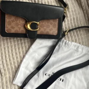 Svart och brun axelväska från Coach - Snygg axelväska från Coach i svart och brun med ett stort guldfärgat spänne. Väskan har ett klassiskt mönster på framsidan och en justerbar axelrem. Insidan är fodrad i brunt tyg med en innerficka för extra förvaring. Den är i toppen skick och man får den med påsen. Köpt från zalando! 