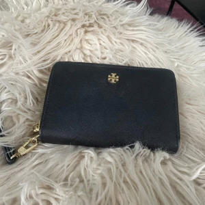 Svart plånbok från Tory Burch - Snygg svart plånbok från Tory Burch med dragkedja och gulddetaljer. Perfekt för att hålla ordning på kort och kontanter. Stilren design med flera fack inuti.