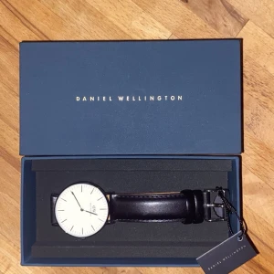 Klocka från Daniel Wellington - Snygg klocka från Daniel Wellington med svart läderarmband och vit urtavla. Klockan har silver detaljer och svart läder rem och kommer i originalförpackning. Knappt använd och nytt batteri i den! Kostar 1999kr i butik