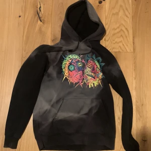 Svart hoodie med färgglatt tryck - Säljer en svart hoodie med ett unikt och färgglatt tryck på framsidan. Hoodien har en stor ficka framtill och en bekväm huva. Perfekt för en cool och avslappnad stil. Pris ej hugget i sten å våga fråga !😁😊( snörena är lite söndriga)