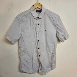 Vit kortärmad skjorta från Tommy Hilfiger - Säljer en stilren vit kortärmad skjorta från Tommy Hilfiger i slim fit. Skjortan har ett diskret prickigt mönster och knappar framtill. Perfekt för en avslappnad men ändå snygg look.