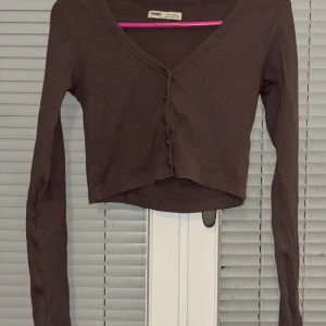 Brun ribbad kofta från Pull&Bear - Säljer en brun ribbad kofta från Pull&Bear. Den har en v-ringad design med knappar framtill och är croppad i modellen. Perfekt för en stilren och avslappnad look.