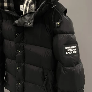 Svart dunjacka från Burberry - Säljer en snygg svart dunjacka från Burberry med märkets logga på ärmen. Jackan har en quiltad design och en avtagbar huva med ett klassiskt rutigt mönster. Perfekt för kalla dagar med sin varma och stilrena look.                                 den går även och göra till väst och sitter jätte bra.kan även tänka mig byta mot en canda gose jacka👍🏼