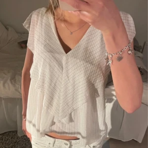 Vit blus med volangdetaljer - Säljer en söt vit blus med subtila ränder och volangdetaljer framtill. Blusen har en v-ringad hals och korta ärmar. 