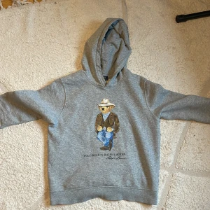 Grå hoodie från Ralph Lauren med björnmotiv - Säljer en grå hoodie från Ralph Lauren med ett coolt björnmotiv på framsidan. Tröjan har långa ärmar och en bekväm huva. Perfekt för en avslappnad stil. 🐻
