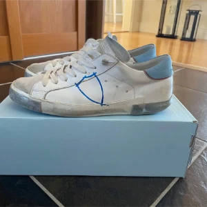Philippe modelle skor - Säljer ett par vita sneakers med blå detaljer och snörning. Skorna har en unik design med en blå linje på sidan och en ljusblå häl. Perfekta för en avslappnad stil. Finns en deefekt vilket är ett hälslitsge på båda skorna och därför erbjuder sänkt pris, är storlek 41 men passar 42 ock