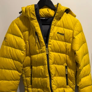 Jacka  - Säljer en snygg gul dunjacka från Bergans of Norway. Jackan har en dragkedja framtill och två praktiska fickor med dragkedjor. Perfekt för kalla dagar med sin varma och vadderade design. Huvan är justerbar för extra skydd mot väder och vind. Sjukt bra pris!