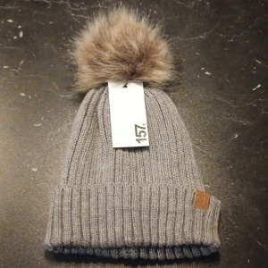 Grå stickad mössa med pälsboll - Snygg grå stickad barn mössa med en stor pälsboll på toppen. Mössan har en ribbad kant och en liten läderdetalj på sidan. Perfekt för kyliga dagar. Den är helt ny och aldrig använd.