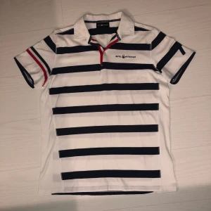 Pikétröja från Sail Racing - Randig pikétröja från Sail Racing. Tröjan är vit med marinblå ränder och har en röd detalj vid knäppningen. Den har korta ärmar med en 7. Vid frågor eller fler bilder kontakta mig. Priset går att diskutera. 