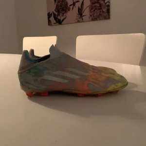 Färgglada fotbollsskor från Adidas - Säljer ett par färgglada fotbollsskor från Adidas med ett unikt mönster i orange, gul och blå. Skorna har en strömlinjeformad design utan snörning och är perfekta för snabba rörelser på planen. De har en låg profil och är designade för att ge bra grepp på gräs.