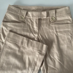 Beige byxor med knappar - Snygga beige byxor med låg midja och vida ben. Byxorna har dekorativa knappar fram och bak, samt fickor med lock. Perfekta för en stilren look. Skrynkliga för tillfället men går att stryka! 