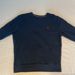 Mörkblå tröja från U.S. Polo Assn. - Säljer en stilren mörkblå tröja från U.S. Polo Assn. med ett rött broderat emblem på bröstet. Tröjan har långa ärmar och en rund halsringning med röd och vit randig detalj på insidan av kragen. NYPRIS: 799 INGA DEFFEKTER