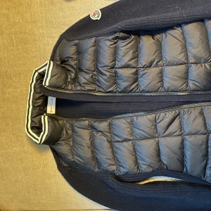 Marinblå dunjacka/kofta från Moncler - Säljer en stilren marinblådunjacka från Moncler. Jackan har en quiltad design med dragkedja framtill och ribbade detaljer vid kragen. Perfekt för kyligare dagar och ger en lyxig känsla. Passar både till vardag och speciella tillfällen. Stl 10 år. Supersnygg och äkta förstås!