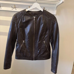 Mörkbrun skinnjacka från Saki Sweden - Snygg mörkbrun skinnjacka från Saki Sweden med dragkedja framtill och två fickor med dragkedjor. Jackan har en stilren design med långa ärmar och quiltade detaljer på axlarna. Perfekt för en cool look! Ej använd endast provad.
