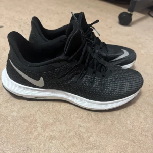 Svarta Nike sneakers - Snygga svarta Nike sneakers med vit sula och klassisk swoosh-logga i vitt. Skorna har snörning och är tillverkade i syntetmaterial, vilket gör dem både lätta och bekväma. Perfekta för träning eller en aktiv vardag.