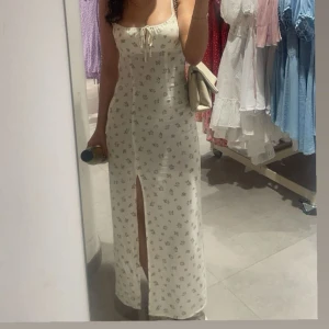 Blommig långklänning med smala axelband - Charmig långklänning i ljus färg med små blommönster. Klänningen har smala axelband och en söt knytning framtill. Perfekt för varma dagar med sin luftiga design.