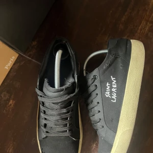 Svarta sneakers från Saint Laurent - Snygga svarta sneakers från Saint Laurent med vit logga på sidan. Skorna har en klassisk design med snörning och en ljus sula som kontrasterar mot det svarta tyget.