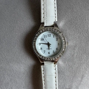 Vit klocka med strass - Säljer en elegant vit klocka med ett glittrande strassbezel. Klockan har en vit urtavla med svarta siffror och visare. Armbandet är i vitt material med en ljus insida, vilket ger en stilren look. Perfekt för både vardag och fest! Märket är Quartz