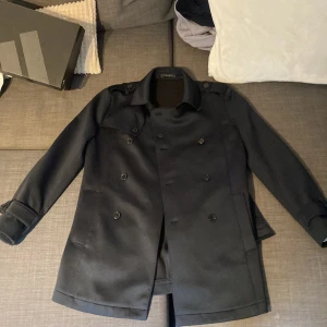 Svart kappa från Zara - Säljer en stilren svart kappa från Zara. Den har en klassisk dubbelknäppt design med knappar och ett bälte i midjan för en snygg passform. Perfekt för höst och vår, och den är långärmad för extra värme. Kappan är i nyskick och redo för nya äventyr!