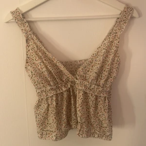 Blommig topp från Shein - Säljer en superfin blommig topp från Shein i storlek S. Den är ärmlös och har ett sött blommönster i olika färger på en ljus bakgrund. Toppen har en smockad detalj på baksidan och en omlott-design framtill. Perfekt för sommardagar!