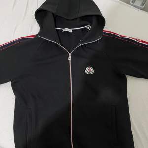 Säljer en snygg svart hoodie från Moncler med dragkedja och huva. Den har röda och vita detaljer längs ärmarna och dragkedjan. Perfekt för en casual look eller när du vill ha något bekvämt men stilrent. Moncler-loggan sitter på bröstet.