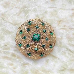 Vintage brosch med gröna stenar - Säljer en superfin vintage brosch i guld med gröna stenar. Okänd metall. Den har ett intrikat mönster med en stor grön sten i mitten och flera mindre runtom. Perfekt för att piffa upp en outfit till fest eller bal. Broschen är cirka 5 cm i diameter.