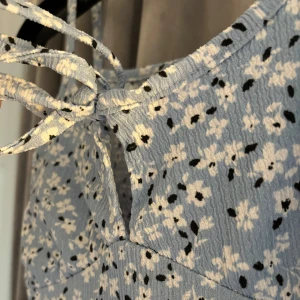 Blommig sommarklänning med knytband - Säljer en superfin blommig klänning i ljusblått med vita och svarta blommor. Den har smala axelband och ett sött knytband framtill. Perfekt för sommardagar och midsommarfirande! 🌸Oanvänd med prislapp kvar!