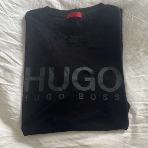 Svart t-shirt från Hugo Boss - Säljer en stilren svart t-shirt från Hugo Boss. Den har en stor logga på framsidan och är perfekt för en casual look. T-shirten är i bra skick och passar till både vardag och fest.