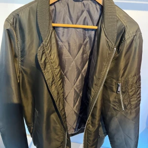 Olivgrön bomberjacka från River Island - Säljer en snygg olivgrön bomberjacka från River Island. Jackan har en quiltad insida och dragkedja framtill. Den är långärmad och har en klassisk bomberstil med ribbade muddar vid ärmslut och krage. Perfekt för höst och vår!