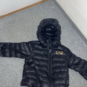 Svart pufferjacka från Emporio Armani - Säljer en snygg svart pufferjacka från EA7 Emporio Armani. Jackan har en huva och dragkedja framtill. Perfekt för kyliga dagar och håller dig varm och stilren. Den är i bra skick och redo för en ny ägare!
