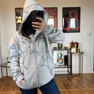 Grå hoodie med mönster - Säljer en supermysig grå hoodie med ett coolt svart mönster. Den har en dragkedja framtill och en stor huva som är perfekt för kyliga dagar. Tröjan är oversized och gjord i mjukt material, vilket gör den perfekt för både vardag och chill. Den har långa ärmar och ribbade muddar vid ärmslut och nederkant. Perfekt för höst och vinter!