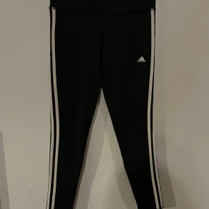 Svarta leggings från Adidas - Säljer ett par svarta leggings från Adidas med vita ränder längs sidorna. De är tillverkade i ett Climalite-material som är perfekt för träning. Leggingsen har en hög midja och en tight passform, vilket gör dem både bekväma och stiliga. Strl S.