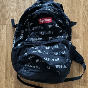 Svart ryggsäck från Supreme - Säljer en  svart ryggsäck från Supreme med deras ikoniska logga tryckt över hela väskan. Den har ett stort huvudfack och flera mindre fack för enkel organisering. Axelremmarna är vadderade för extra komfort. Perfekt för skolan eller vardagsbruk!