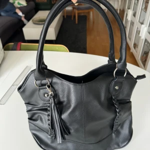 Svart axelväska med flätade detaljer - Snygg svart axelväska i syntetmaterial. Väskan har flätade detaljer på framsidan. Den är rymlig och perfekt för vardagsbruk. Inuti finns det gott om plats för alla dina grejer. Perfekt för både skola och jobb! Hämtas på Hägerstensåsen eller ök 🖤👜