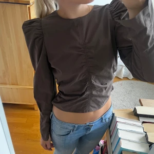 Brun långärmad topp - Säljer en snygg brun långärmad topp med puffiga ärmar och rynkdetaljer framtill. Toppen har en tight passform och är perfekt för höst och vår. Den har en dragkedja vid ärmsluten för en extra touch. Perfekt för både vardag och fest!