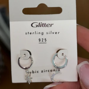 Silverörhängen med stjärna och måne från Glitter - Säljer ett par superfina örhängen i sterling silver från Glitter. Ena örhänget har en hängande stjärna och det andra en måne, båda dekorerade med cubic zirconia för extra glans. Perfekta för att ge en touch av magi till din outfit!