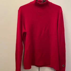 Röd stickad tröja från Busne - Säljer en snygg röd stickad tröja från Busne, tillverkad i Frankrike. Tröjan har en hög krage och långa ärmar, perfekt för kyligare dagar. Den är i ett klassiskt snitt med en liten broderad detalj på ärmen. Passar både till vardags och lite finare tillfällen.