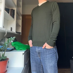 Grön ullkofta från Fred Perry - Säljer en snygg grön tröja från Fred Perry i 100% merinoull. Den har en klassisk v-ringning och långa ärmar, perfekt för höst och vinter. Tröjan är i mycket bra skick och passar både till vardags och lite finare tillfällen. Supermjuk och bekväm!